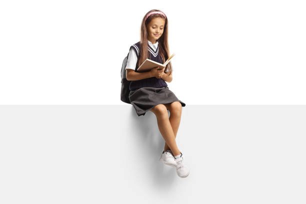 Girl in a school uniform sitting on a blank panel and reading a book isolated on white background