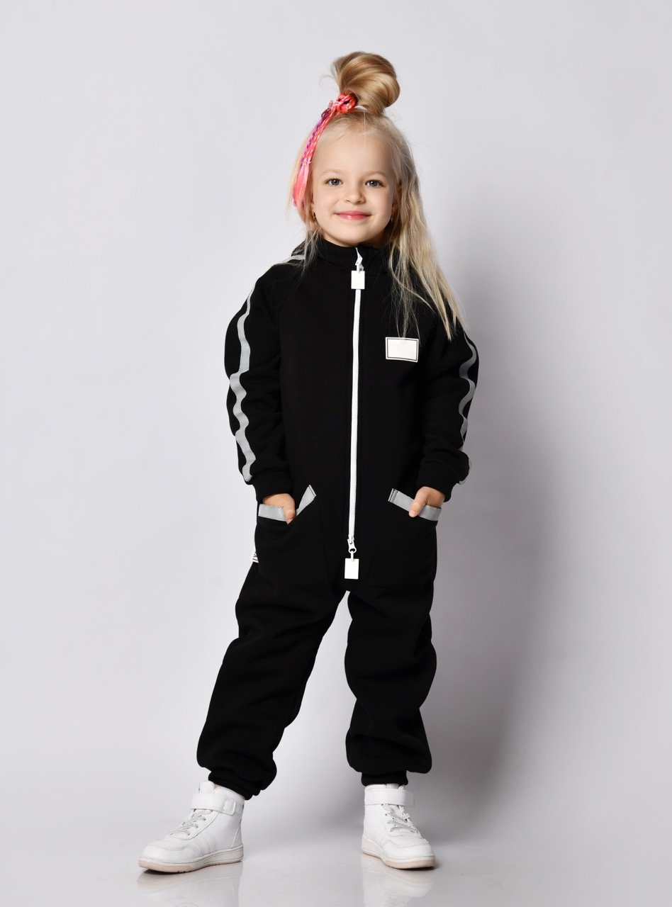 Happy smiling blonde kid baby girl with modern hairstyle and colorful braids in stylish comfortable black jumpsuit and sneakers stands with her hands in pockets over gray background